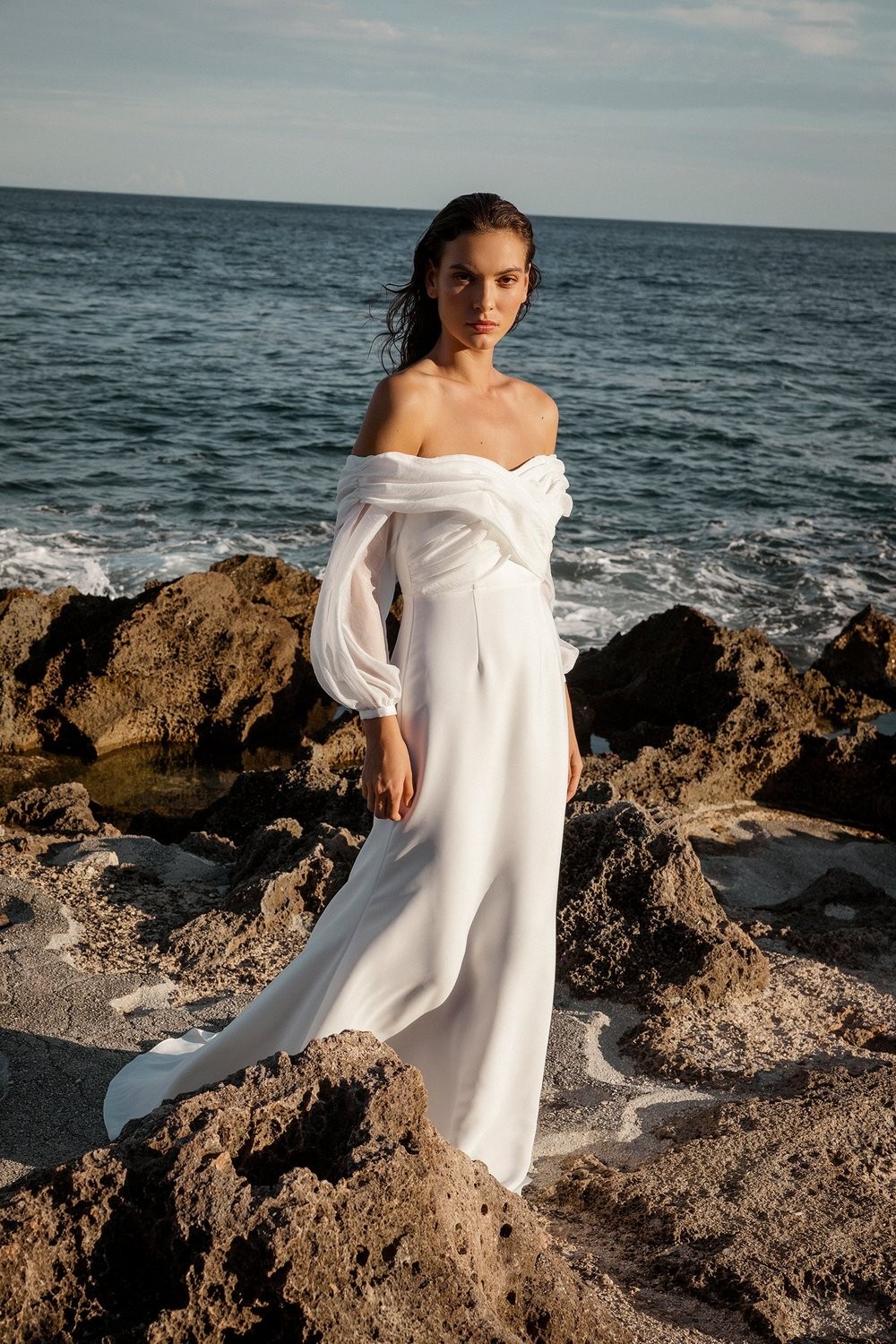 Model by the ocean on cliffs at sunset, wearing a white off-shoulder, long-sleeve wedding dress