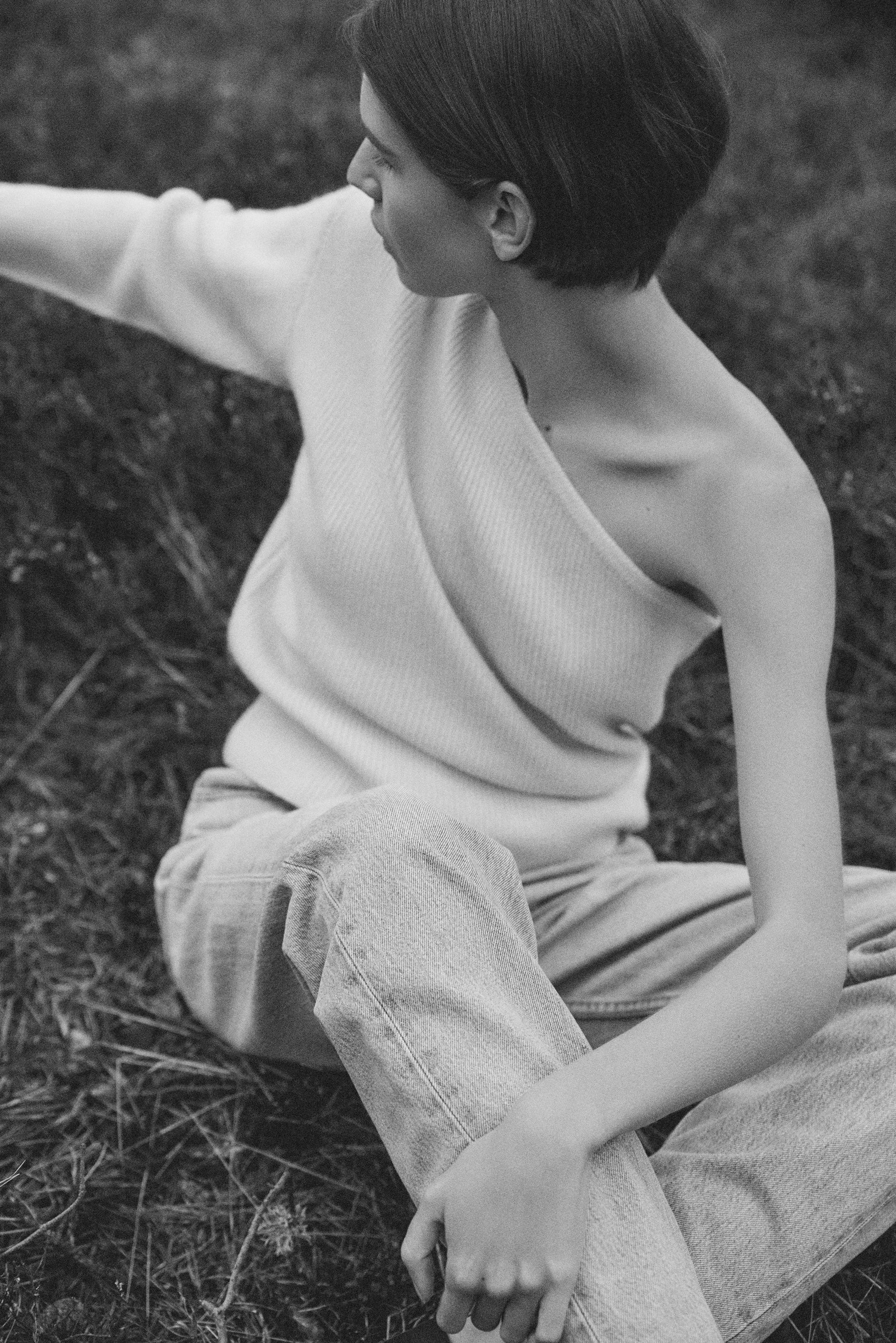 Black and white photo of a model sitting in grass, wearing a one-sleeve shirt and jeans, looking off-camera.